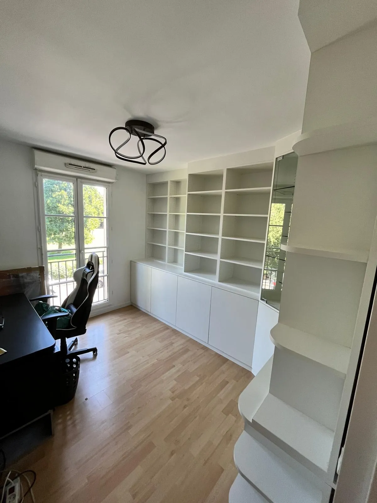 Bureau moderne avec chaise de bureau, large bibliothèque blanche vide et parquet clair près d'une fenêtre avec vue sur des arbres.