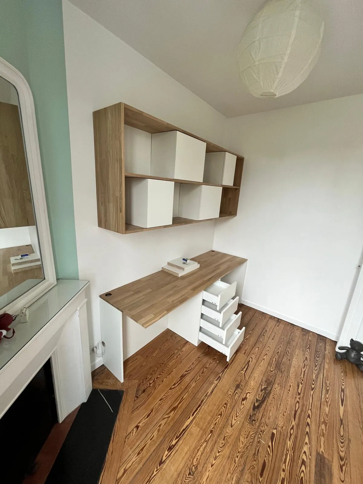 Bureau avec plateau en bois clair et trois tiroirs ouverts, étagères murales avec cubes blancs, sol en parquet et murs blancs.