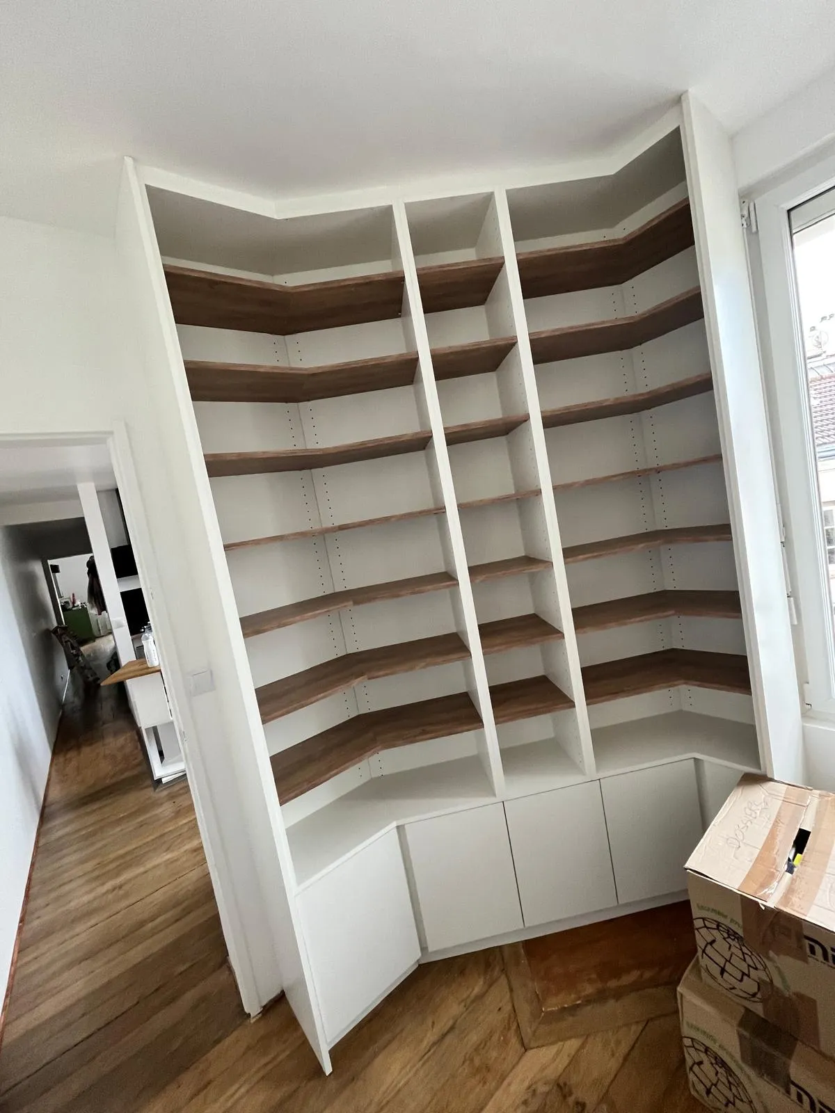 Étagères d'angle en bois avec base blanche et compartiments ouverts dans un coin de pièce lumineuse avec sol en bois.