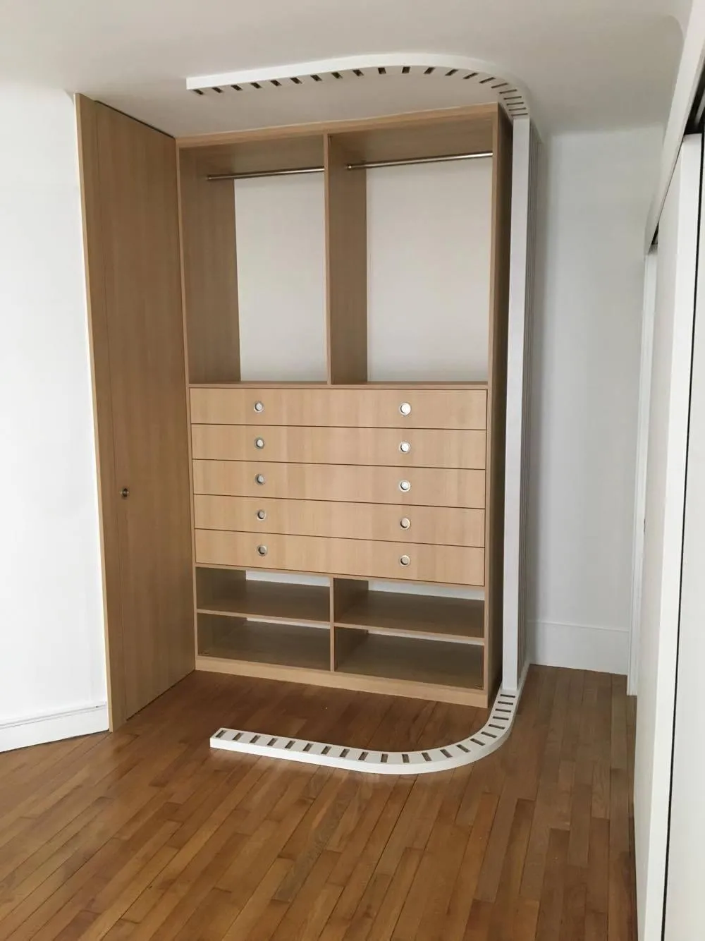 Armoire en bois clair avec tiroirs, étagères et barres pour suspendre des vêtements, avec un conduit d'aération blanc incurvé au plafond et au sol.