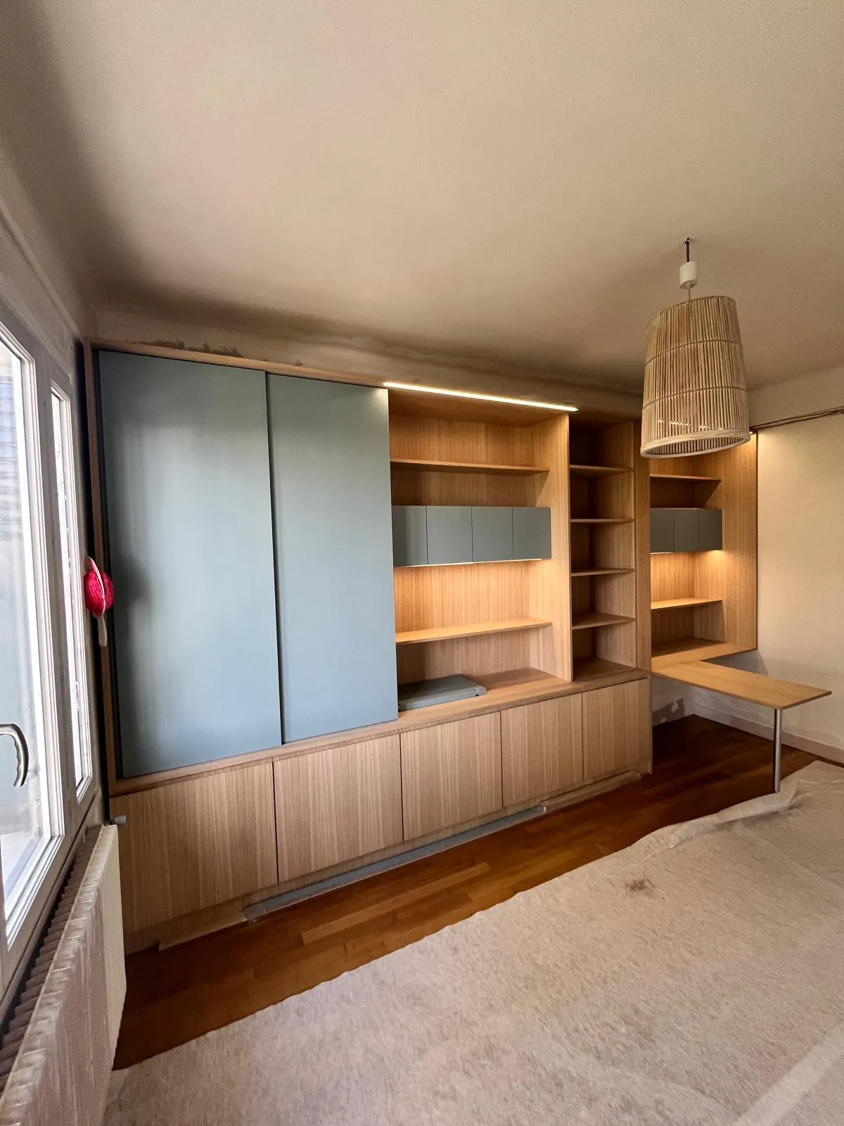 Chambre avec grands meubles en bois clair intégrant des armoires bleues, étagères ouvertes, bureau rabattable et un plafond avec suspension en rotin.
