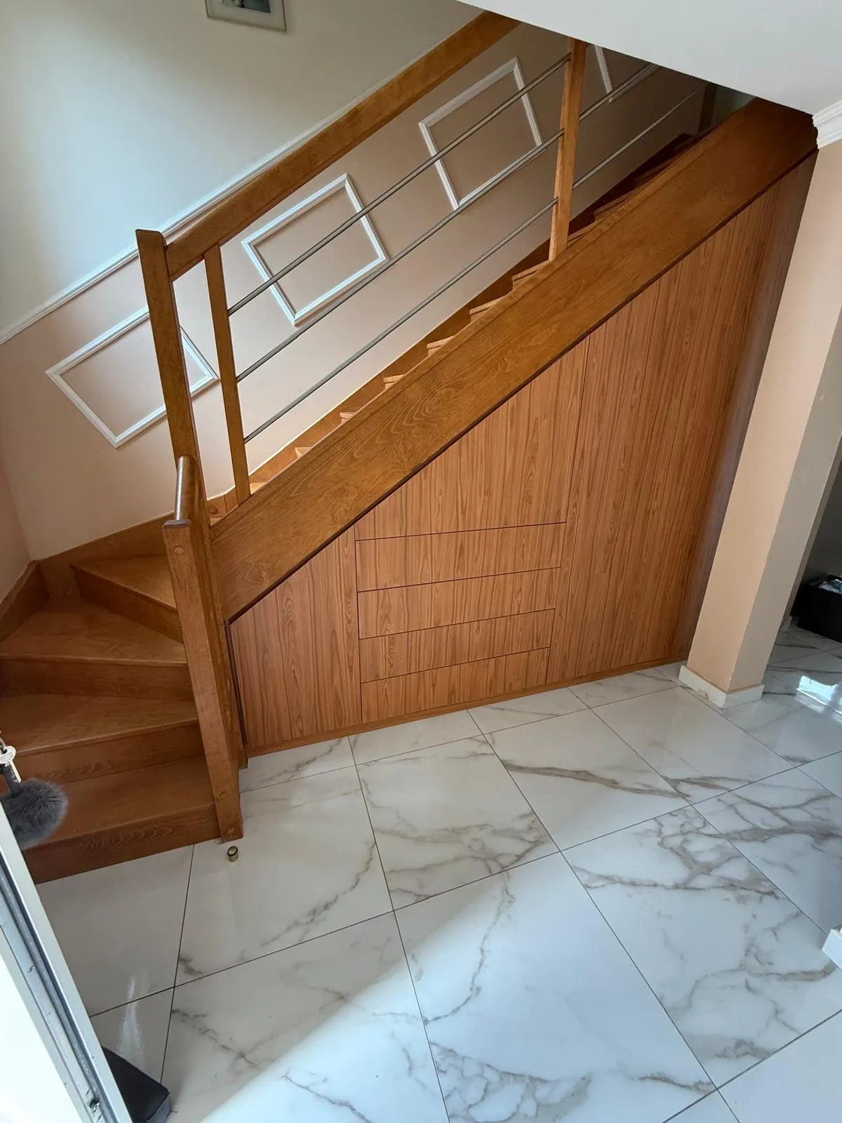 Escalier en bois avec rambarde métallique au-dessus d'un espace de rangement intégré sous les marches, et sol en carrelage blanc marbré.