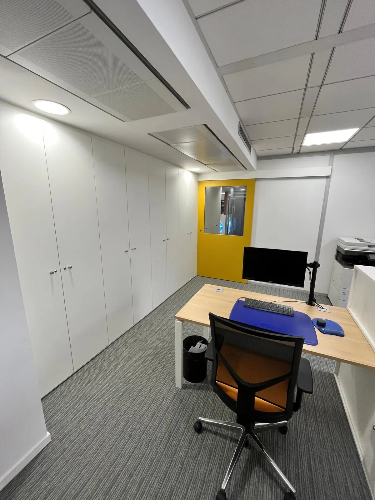 Bureau moderne avec chaise à roulettes, ordinateur sur un bureau en bois clair, grandes armoires blanches et porte jaune avec une fenêtre.