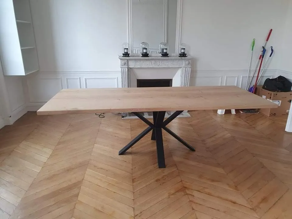 Table en bois clair avec un pied central en métal noir en forme d'étoile, dans une pièce avec parquet en chevrons et une cheminée blanche ornée de quatre lanternes.
