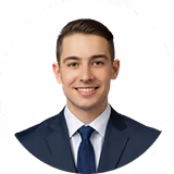 Smiling young man wearing a dark suit and blue tie against white background.
