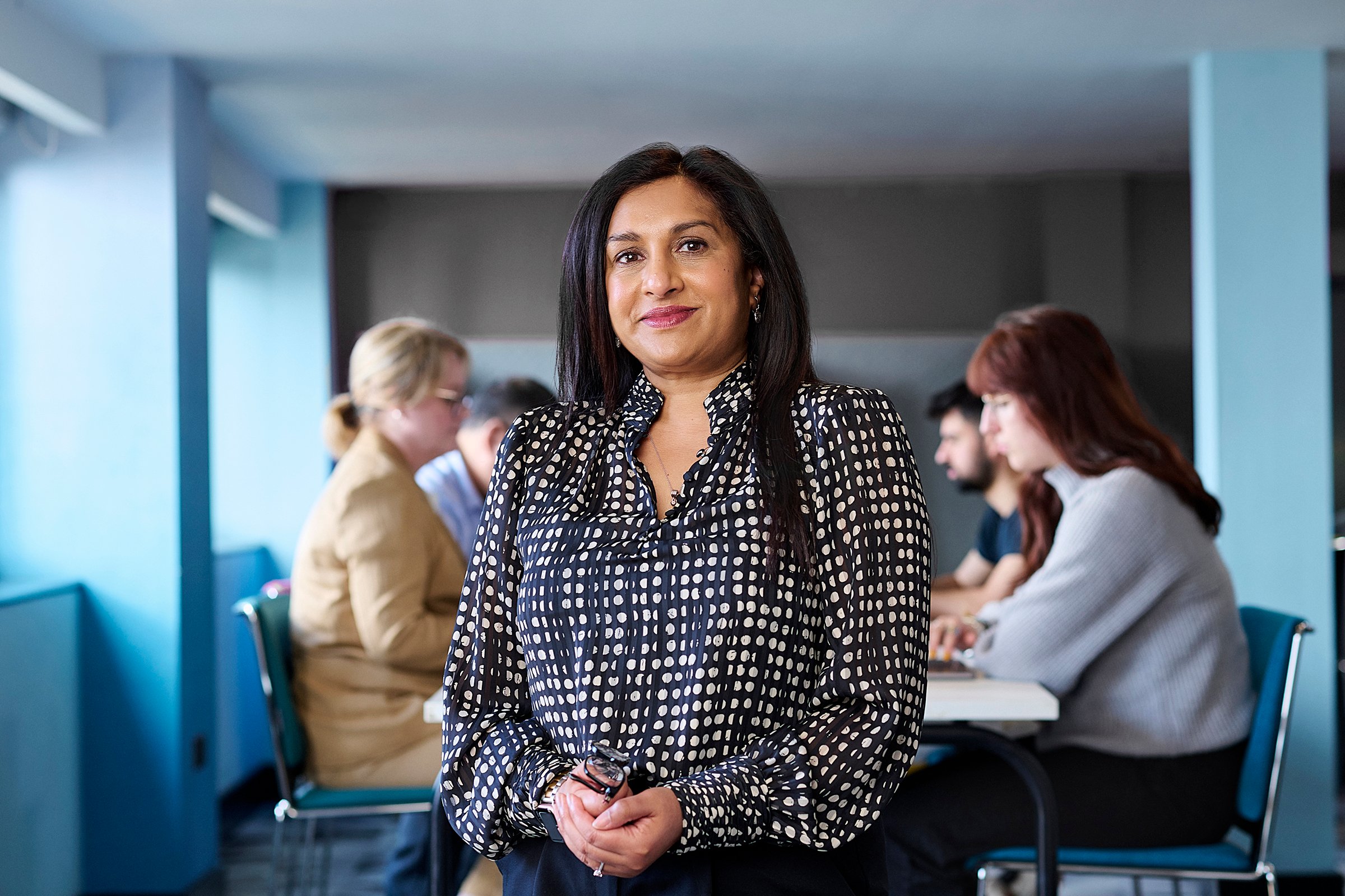 Confident woman standing in an office with colleagues working at a table in the background.