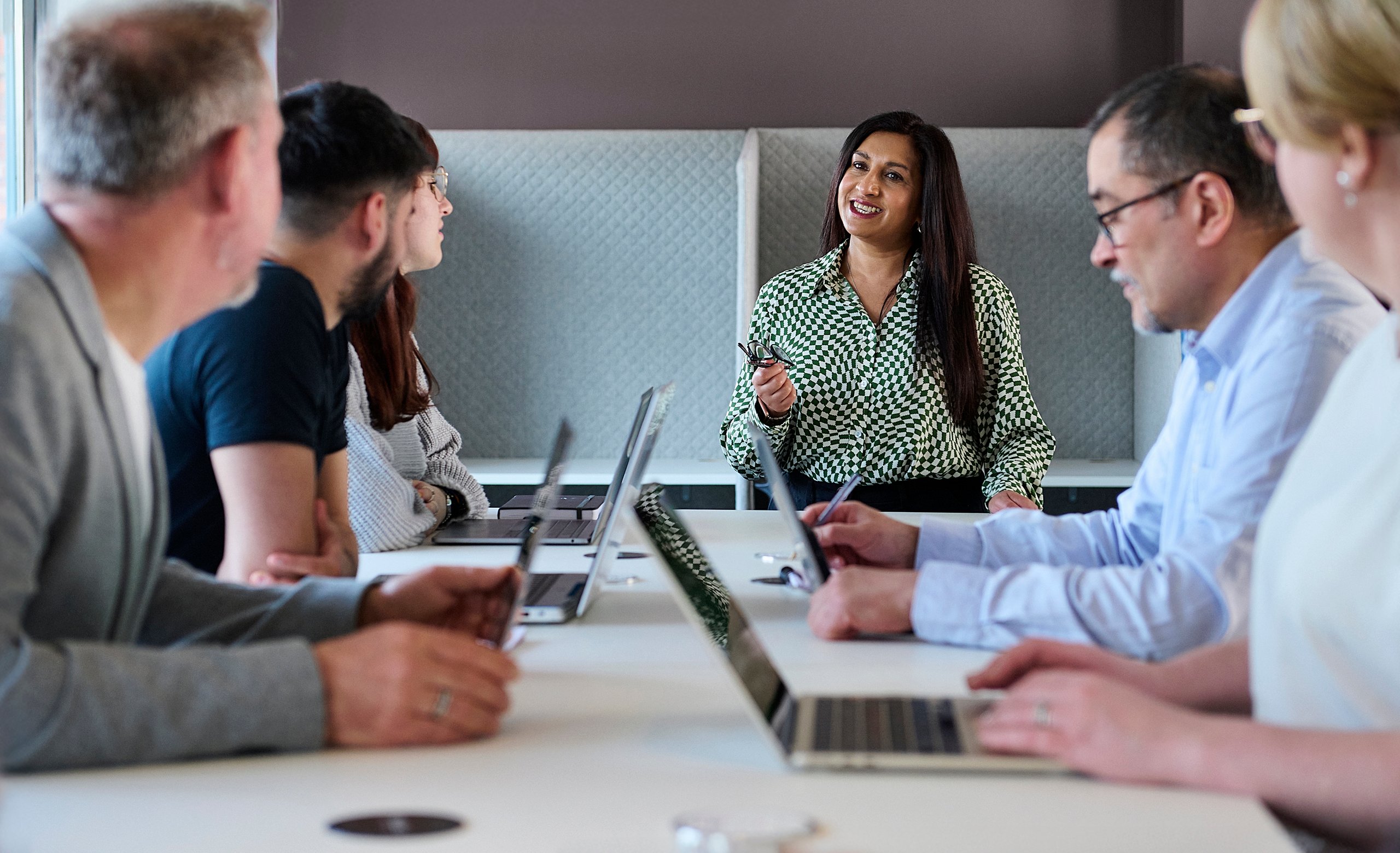 A diverse group of five people sitting around a table with laptops, listening to a woman speaking and smiling.