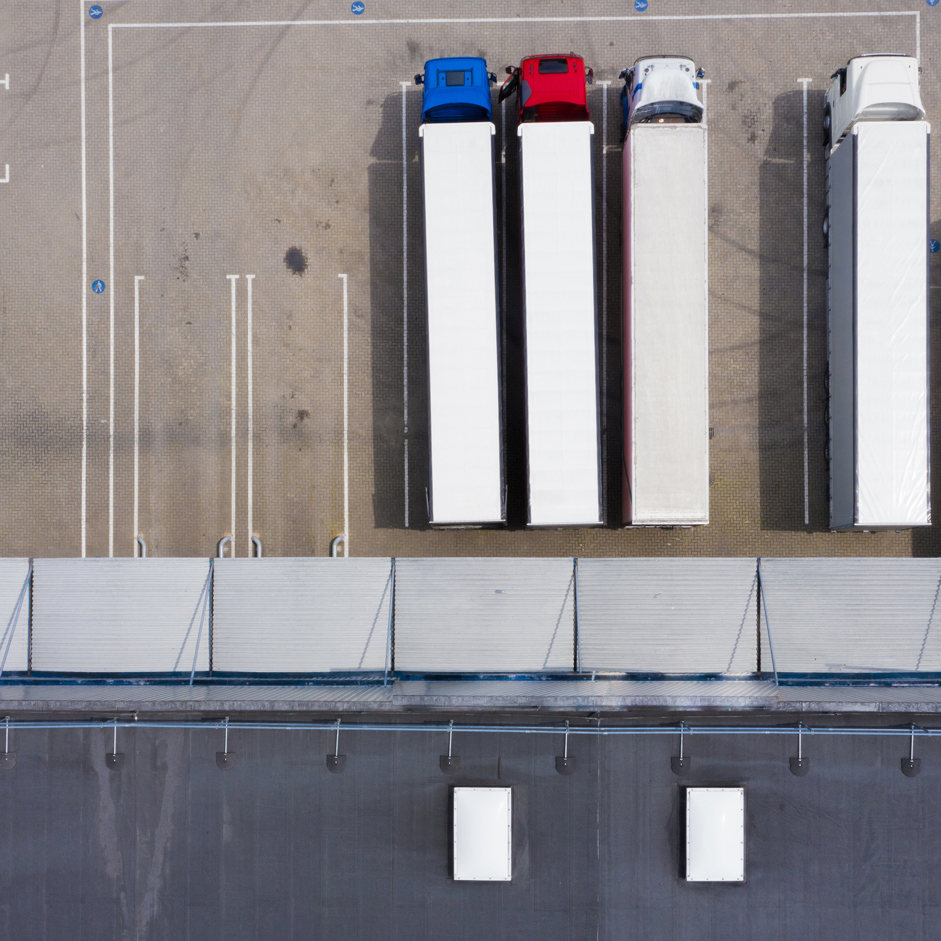 Aerial view of four parked semi-trailer trucks in a loading dock area with a paved surface and pedestrian walkway markings.