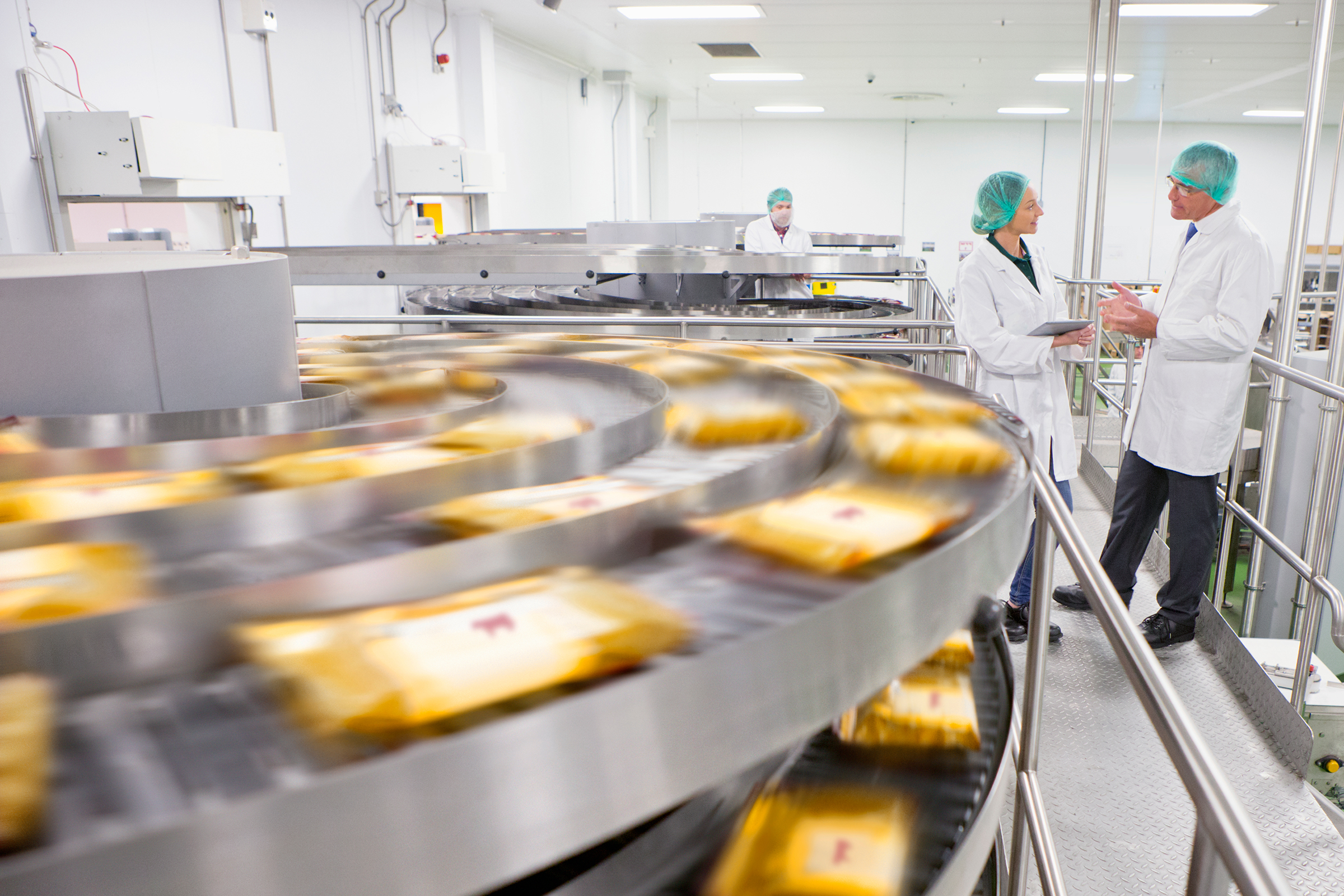 Two factory workers in white coats and hairnets discussing near a conveyor belt with packaged products in a food processing facility.