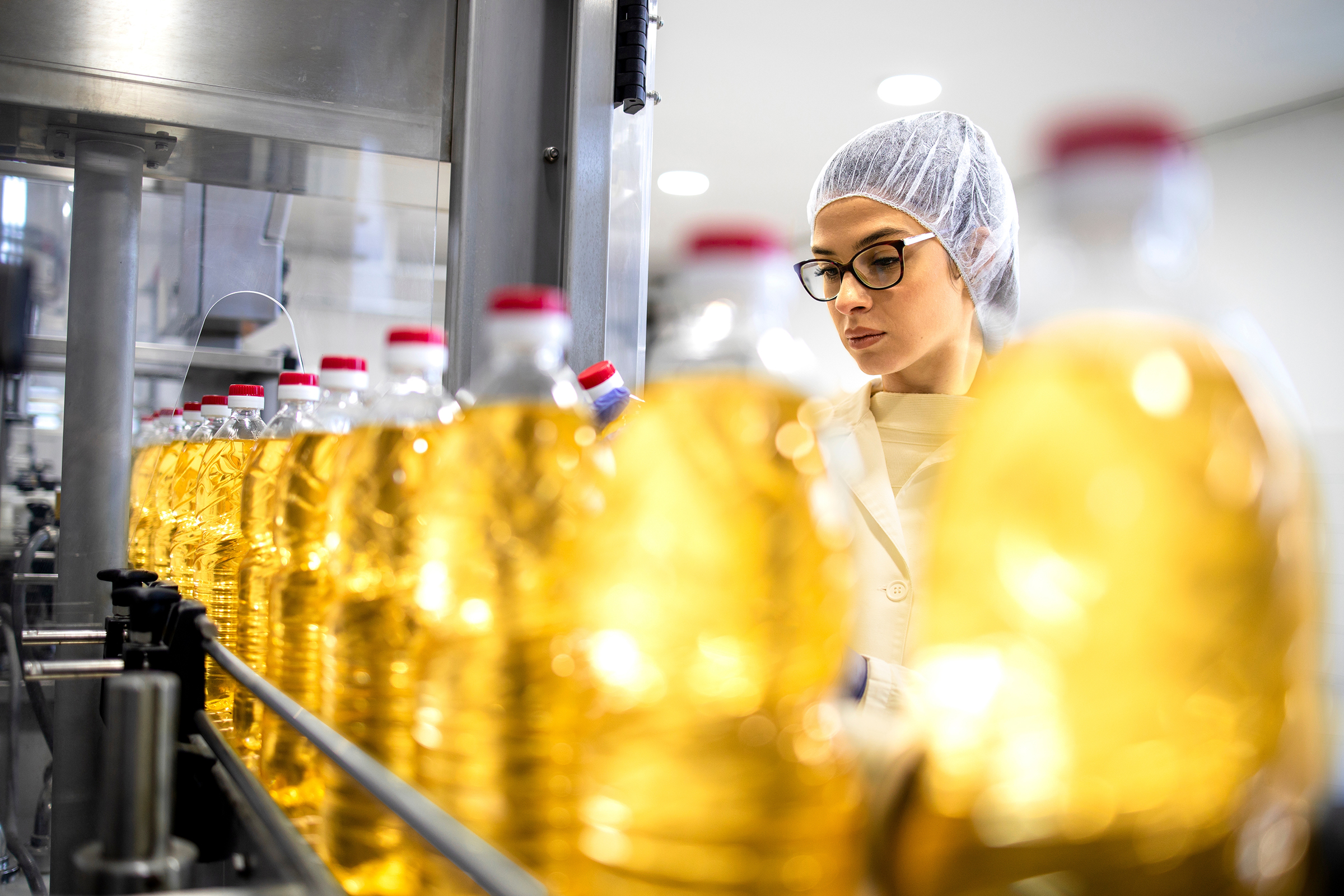 Woman wearing hairnet and glasses inspecting bottles of cooking oil on a production line.