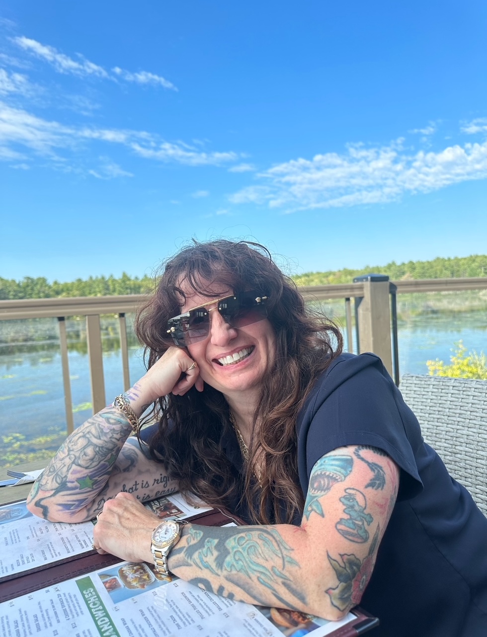 Smiling woman with long curly hair, wearing sunglasses, showing colorful tattoos on both arms, sitting at an outdoor table with menus against a lake and blue sky background.