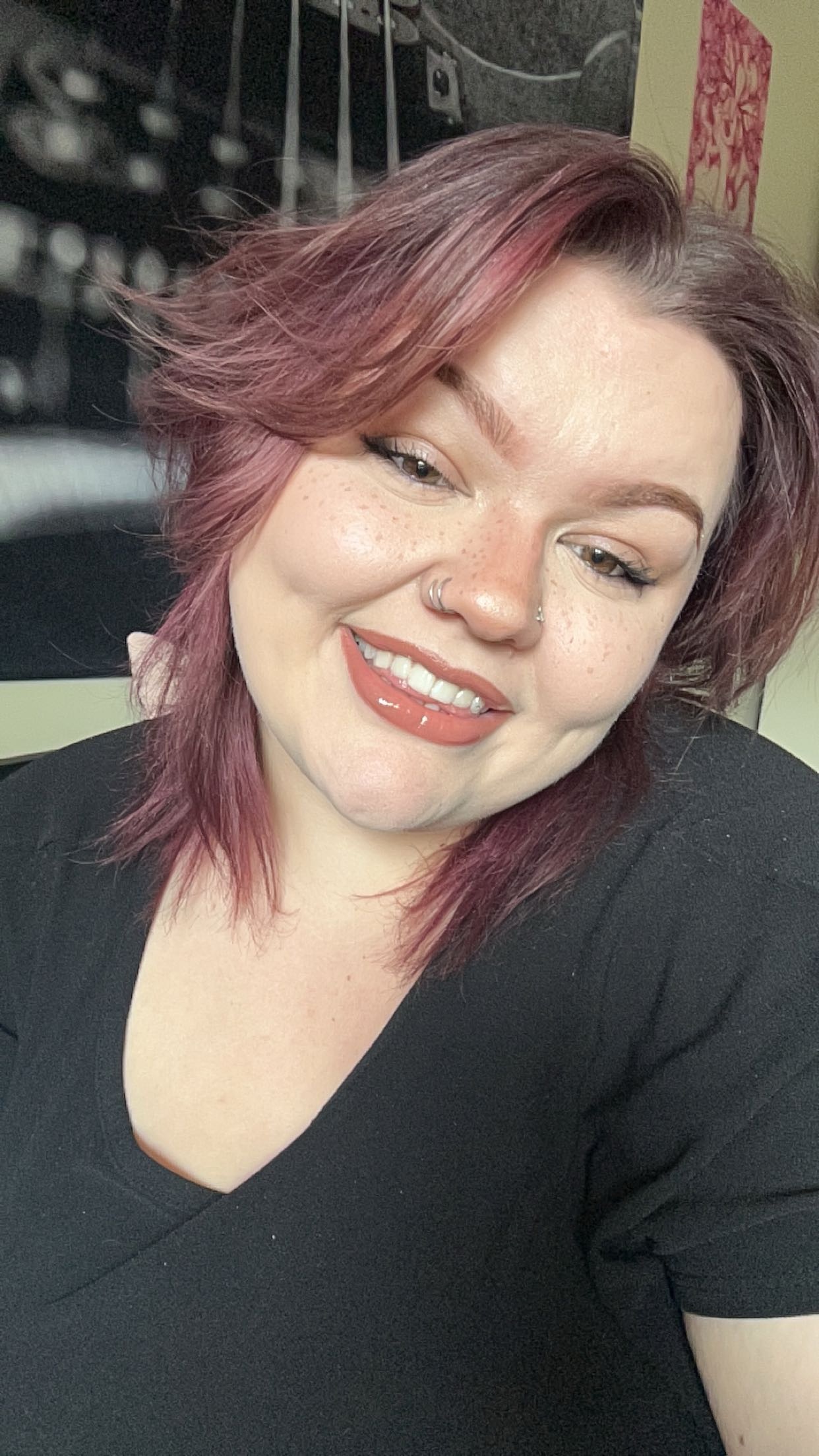 Smiling woman with pinkish-purple hair, freckles, and nose rings wearing a black top.