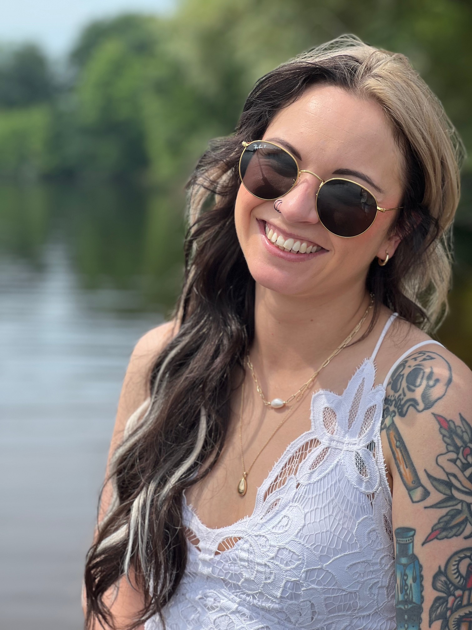 Smiling woman with long dark and blonde hair, wearing round sunglasses, a white lace top, and showing tattoos on her arm, standing by a body of water with trees in the background.