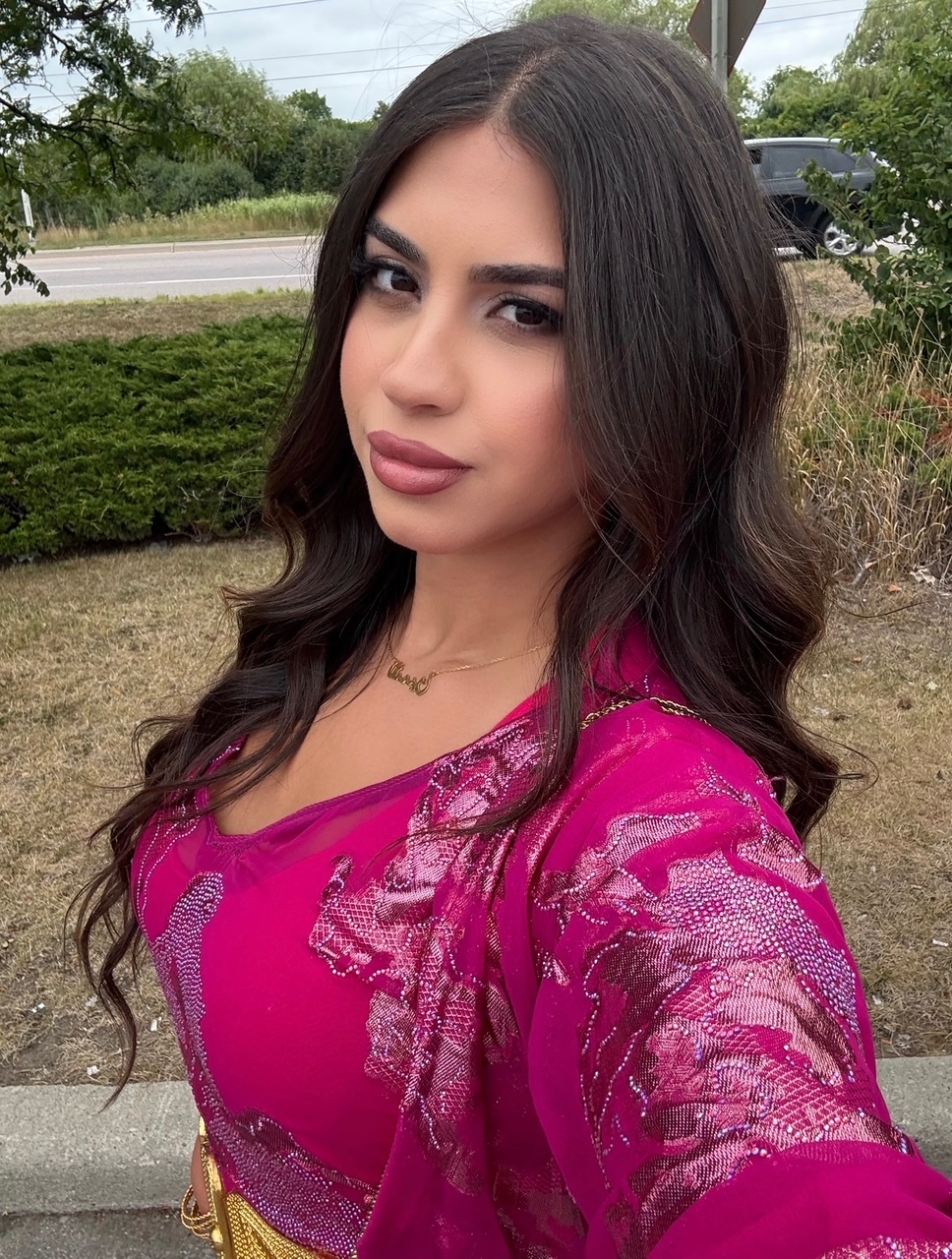 Woman with long dark hair wearing a bright pink traditional outfit with intricate patterns, outdoors near greenery and a road.