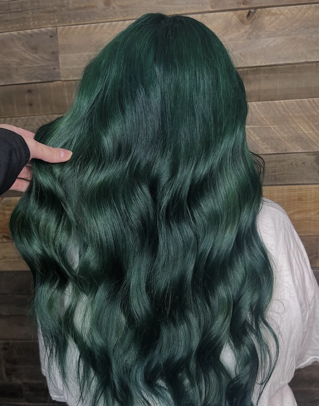 Long, wavy hair dyed a deep green color being held by a hand against a wooden background.