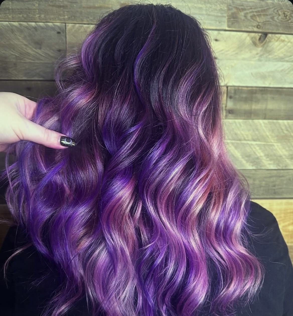 Back view of long wavy hair dyed in vibrant shades of purple with pink highlights against a wooden wall.