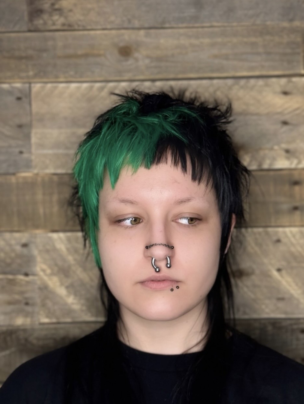 Person with short, two-tone green and black hair, multiple nose and lip piercings, wearing a black shirt against a wooden plank background.