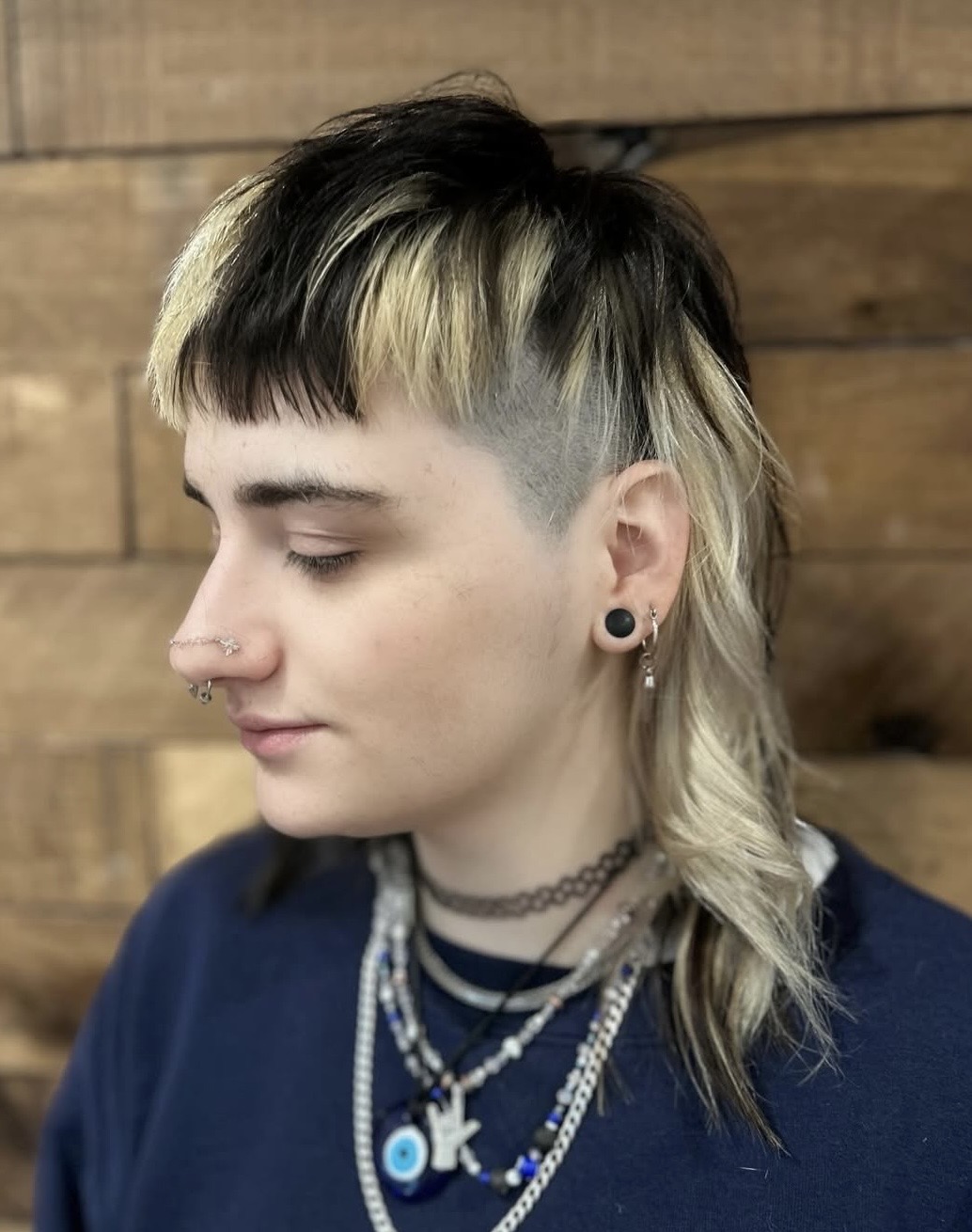 Person with a black and blonde mullet hairstyle, multiple necklaces, and facial piercings, wearing a dark blue shirt against a wooden background.
