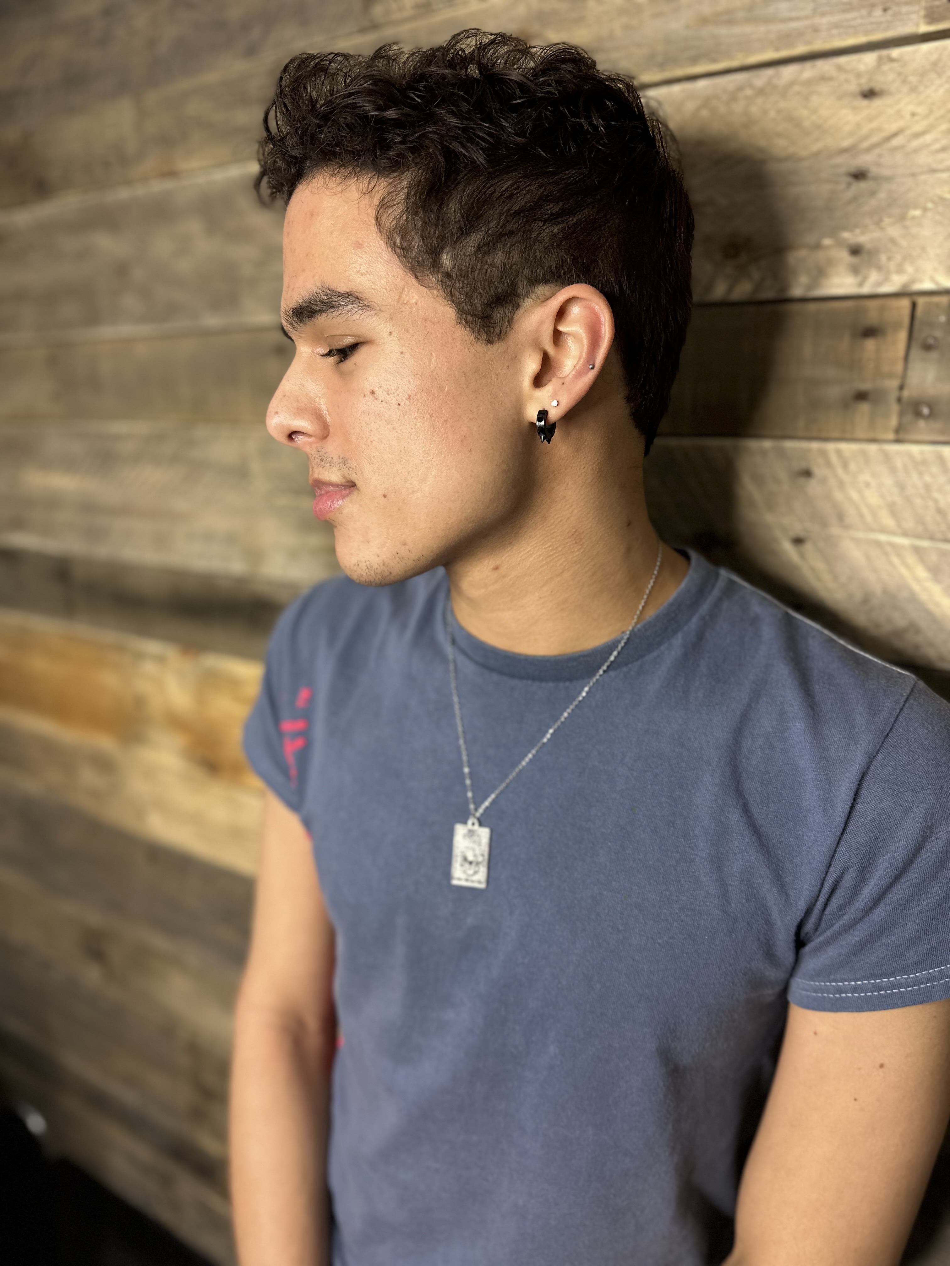 Young man with short curly hair, wearing a blue t-shirt and silver necklace, standing against a wooden wall looking to the side.