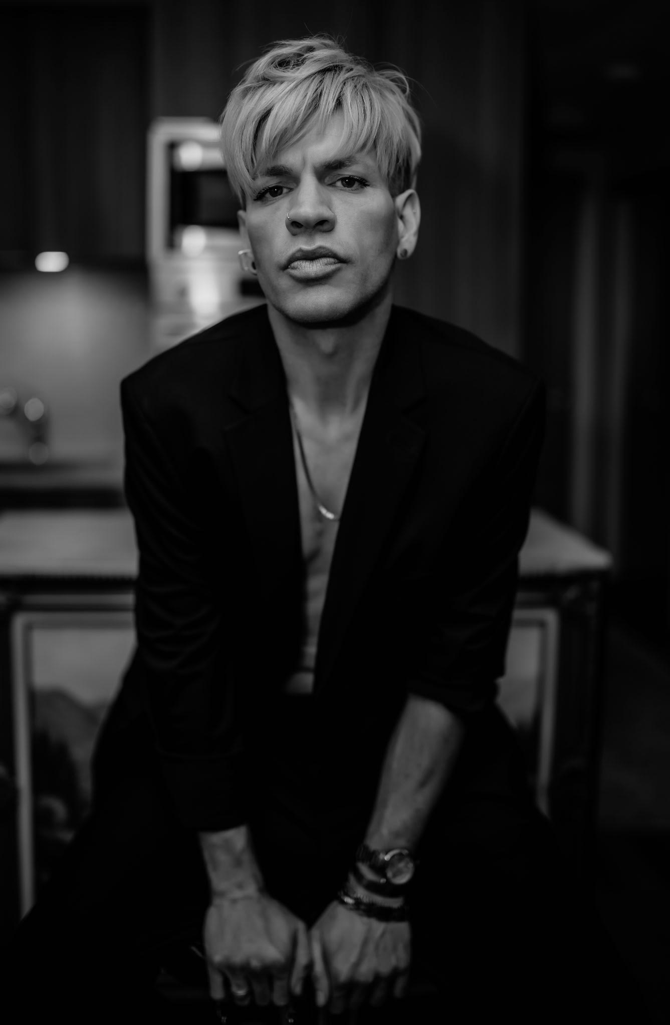 Black-and-white portrait of a person with short light-colored hair and nose piercing wearing a dark blazer, sitting and looking directly at the camera.