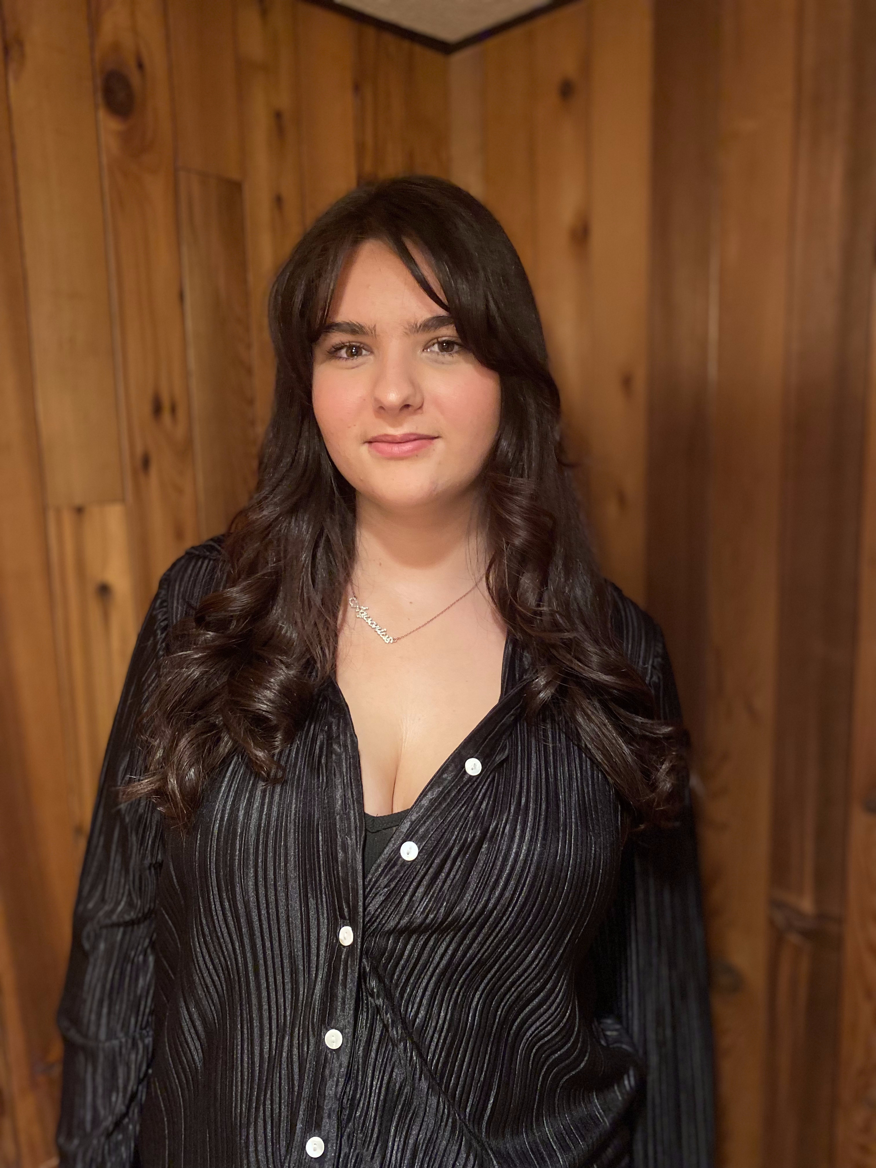 Young woman with long dark wavy hair wearing a black pleated blouse and a necklace, standing in front of a wooden wall.