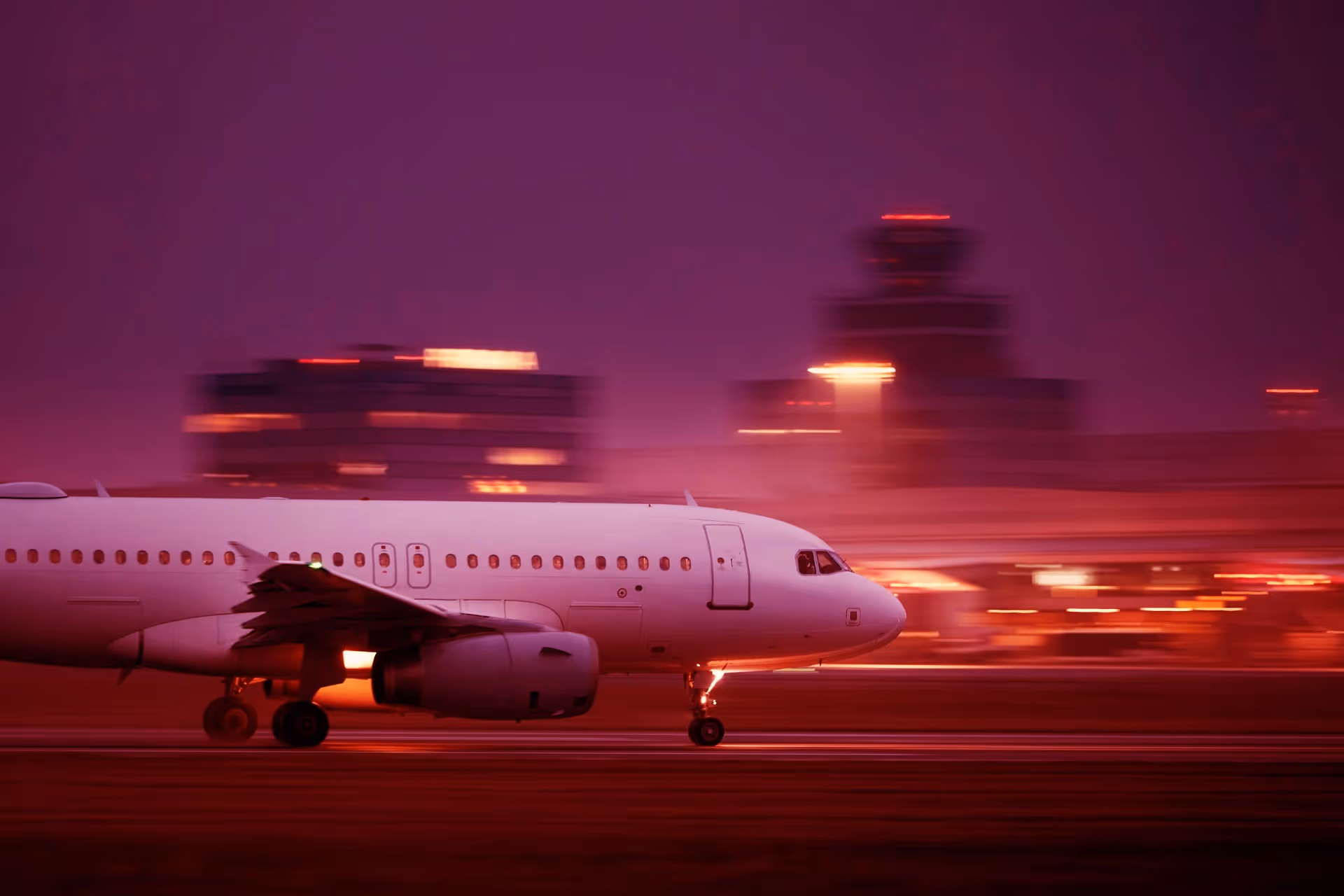 White airplane taxiing on runway at dusk with blurred airport buildings and lights in background.
