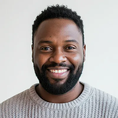 Smiling man with short curly hair and beard wearing a gray knit sweater against a plain light background.