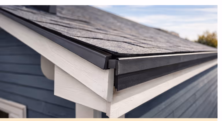 Close-up of a roof corner with gray asphalt shingles and black gutter system on a blue house with white trim.