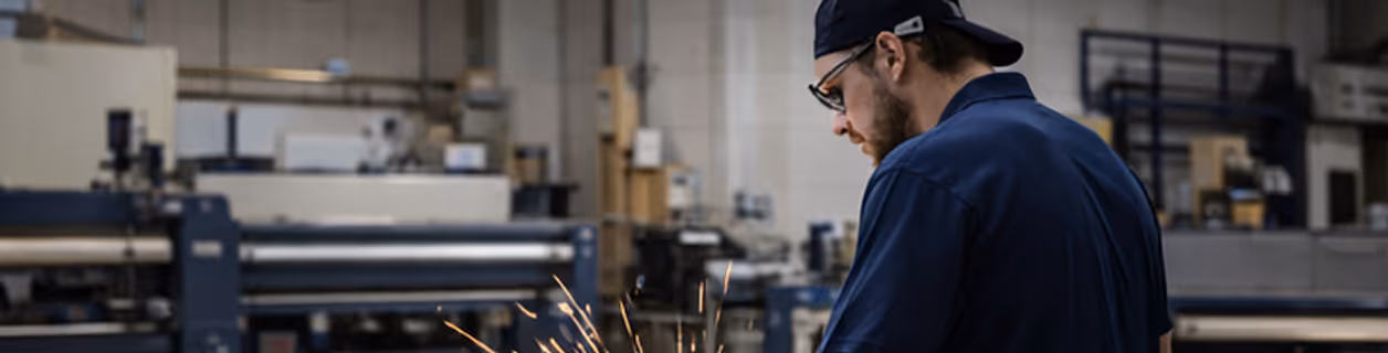 Man wearing safety glasses and a cap using a grinder tool that emits sparks in a workshop.