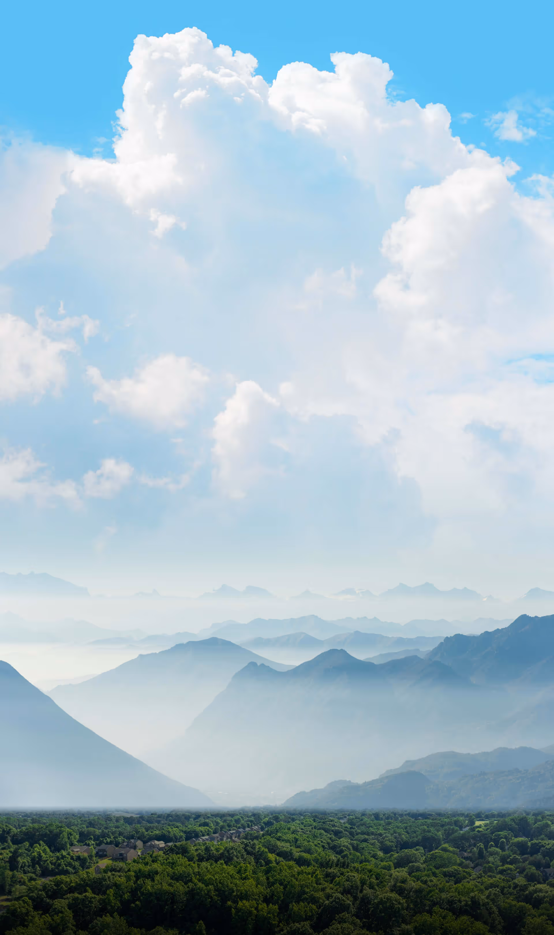 Layered mountain ranges shrouded in mist over a green forest under a blue sky with large fluffy clouds.