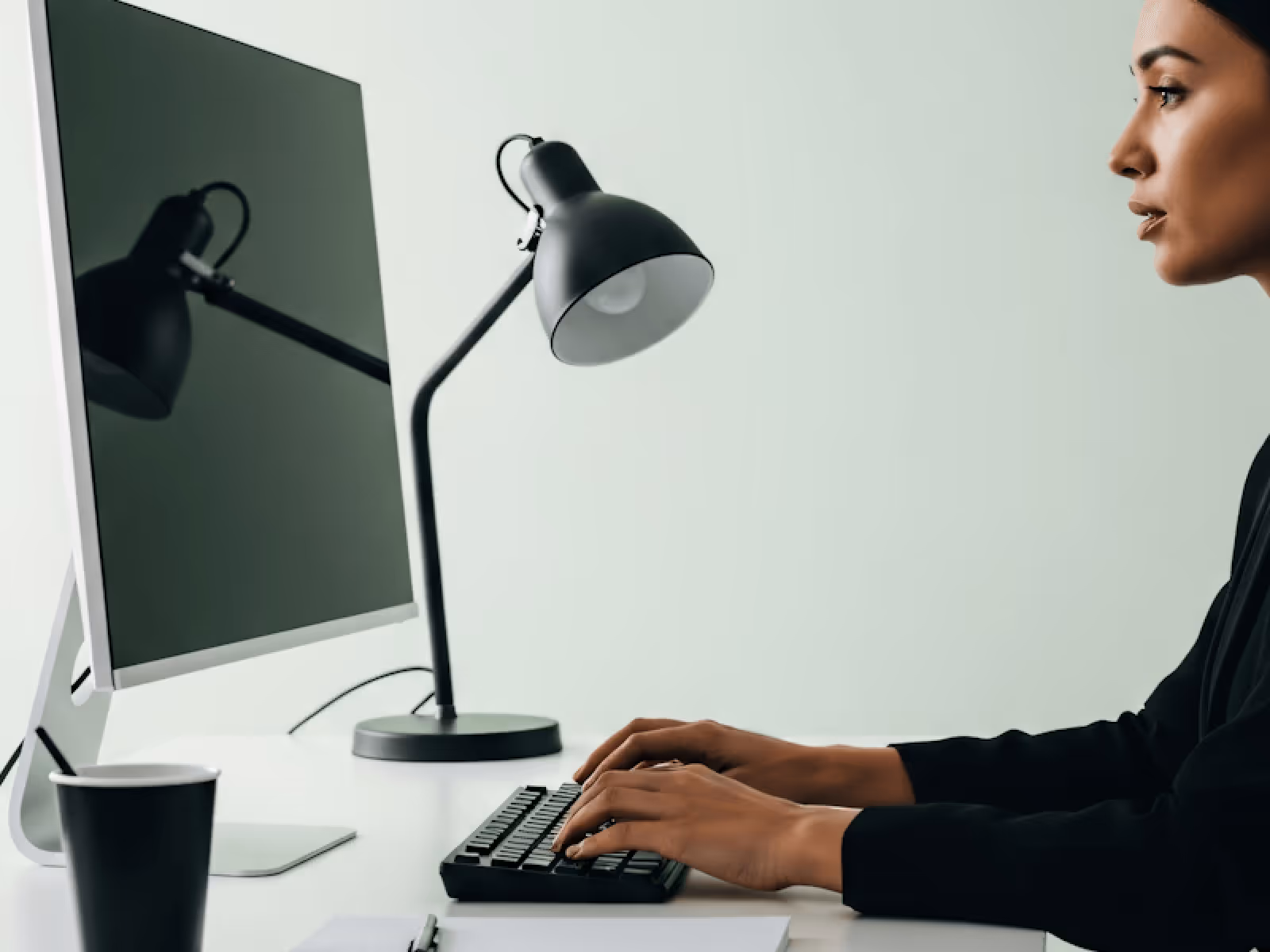 Side view of a woman typing on a keyboard at a desk with a monitor, desk lamp, and a black coffee cup.