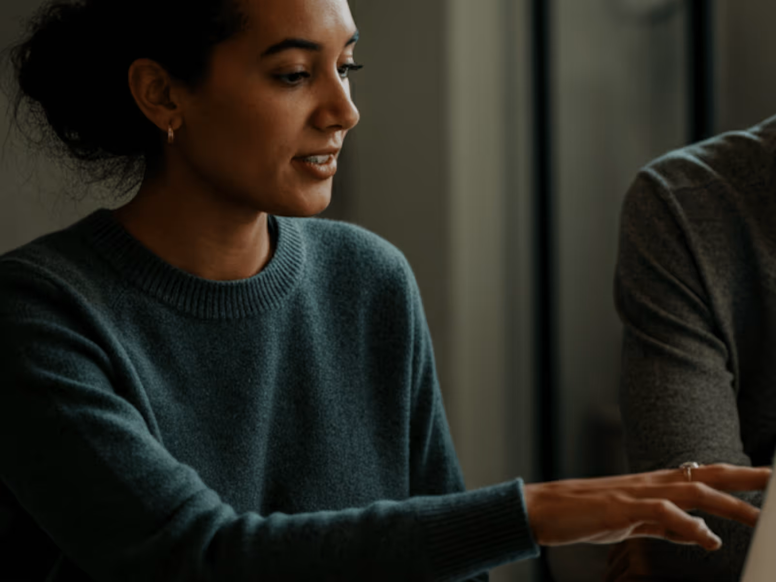 A woman in a green sweater is pointing at something on a laptop screen while talking to another person.