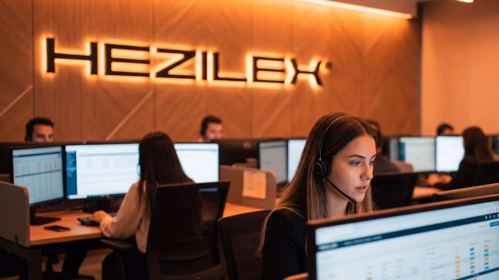 Mulher com headset trabalhando em um escritório moderno com vários monitores e o letreiro iluminado 'HEZILEX' na parede.