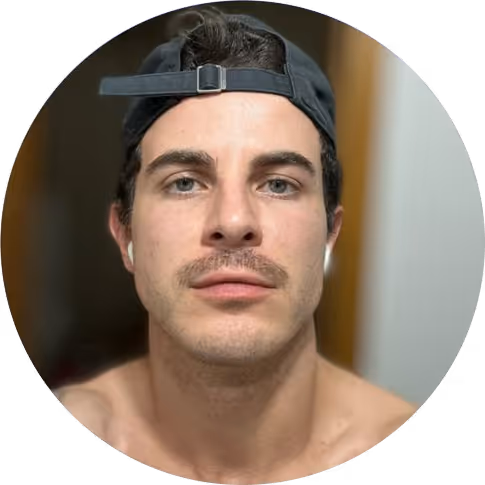 Homem jovem com boné preto virado para trás e fones de ouvido brancos, olhando diretamente para a câmera.