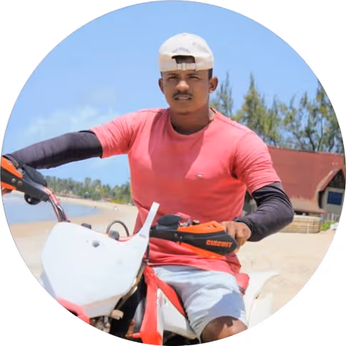 Homem usando boné branco e camiseta vermelha sentado em uma moto branca na praia.