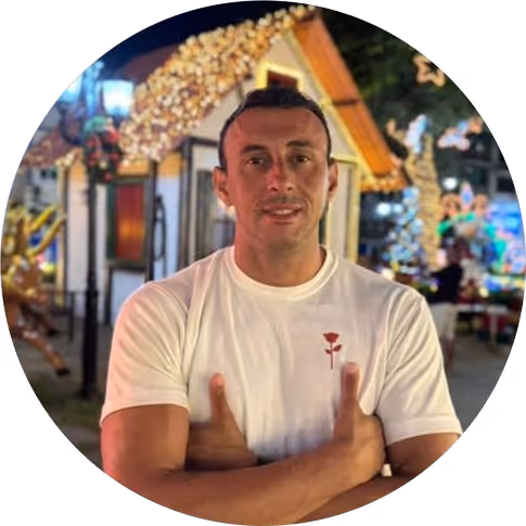 Homem de camiseta branca com desenho de rosa, braços cruzados, em ambiente externo iluminado com decoração de luzes ao fundo.