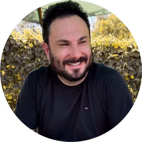 Homem sorridente com barba e cabelo preto, vestindo camiseta preta, sentado ao ar livre com vegetação ao fundo.