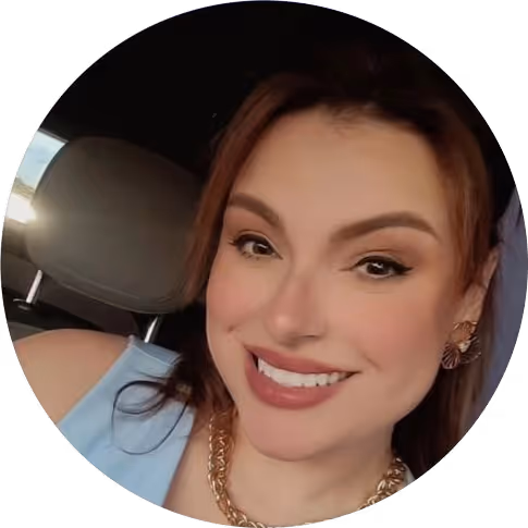 Mulher sorridente com maquiagem suave, usando brincos e colar dourado, vestindo uma blusa azul clara dentro de um carro.