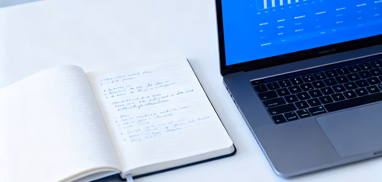 Open notebook with handwritten notes beside a laptop displaying a blue screen with charts and data.