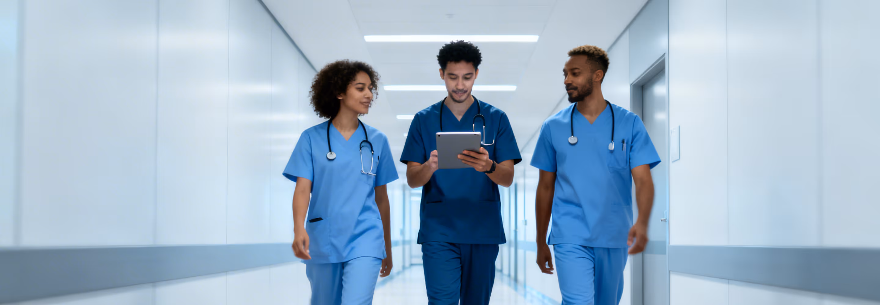 Three doctors in blue scrubs walking down a hospital corridor, one holding a tablet.