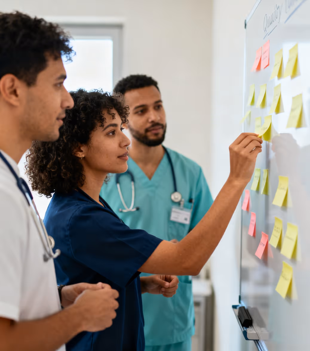 Three healthcare professionals discuss and arrange sticky notes on a whiteboard during a meeting.
