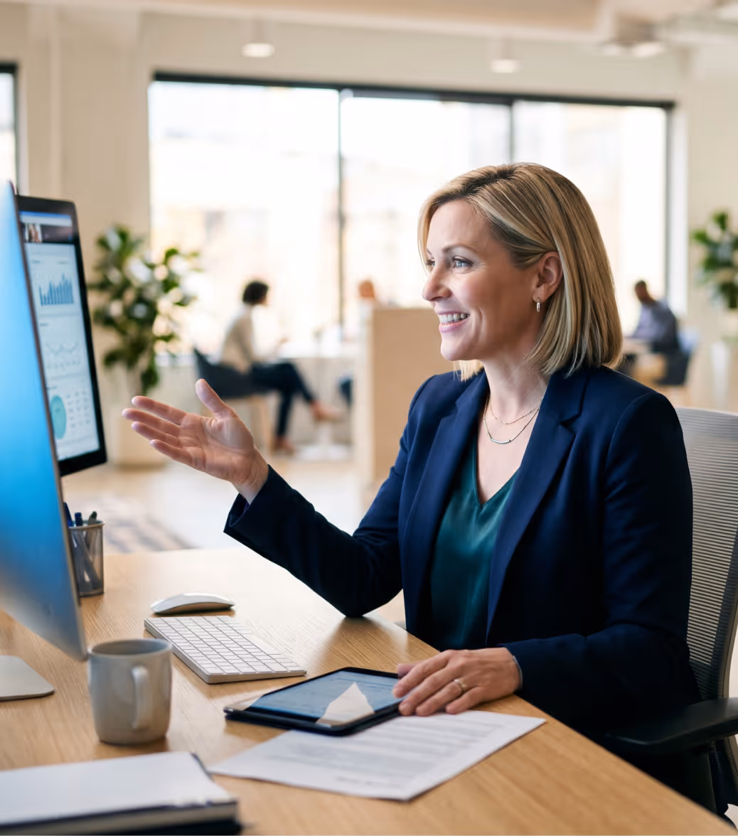 Smiling businesswoman gesturing while working at a desk with computer and tablet in office.