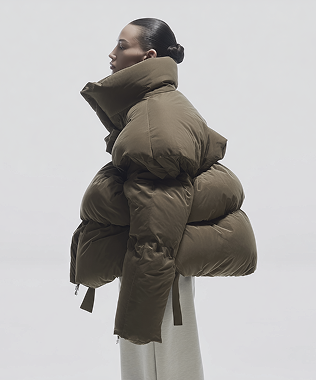 Side profile of a person wearing an oversized olive green puffer jacket with a high collar and light-colored pants against a plain background.