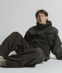 Young person sitting on the floor wearing a black jacket and black pants against a plain light gray background.
