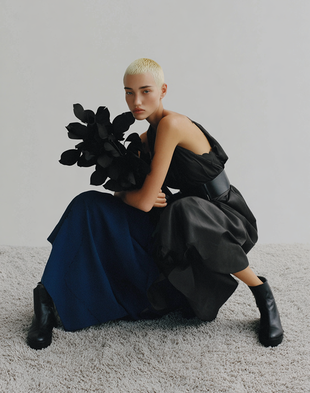 Person with platinum blonde short hair wearing a black dress and boots, sitting on a carpeted floor holding black paper leaves.