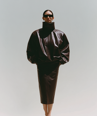 Person wearing oversized black leather jacket and matching pencil skirt with sunglasses against a plain background.