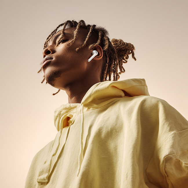 Young man with dreadlocks wearing a yellow hoodie and wireless earbuds looking upwards against a soft gradient background.