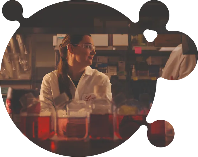 A female scientist in a white lab coat and safety goggles standing among laboratory containers filled with red liquid in a warmly lit lab.