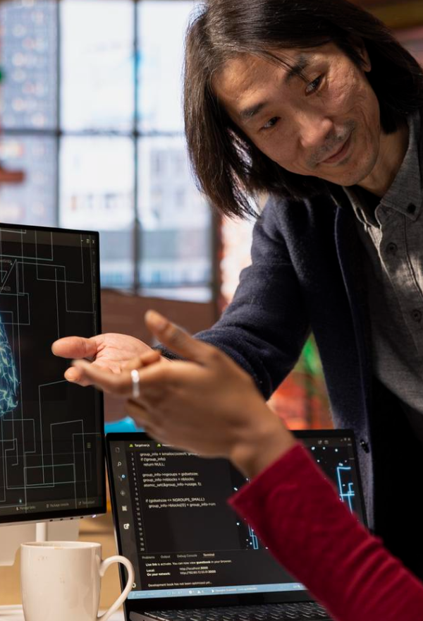 Two men standing closely, looking down and engaging in discussion near a sunlit window.