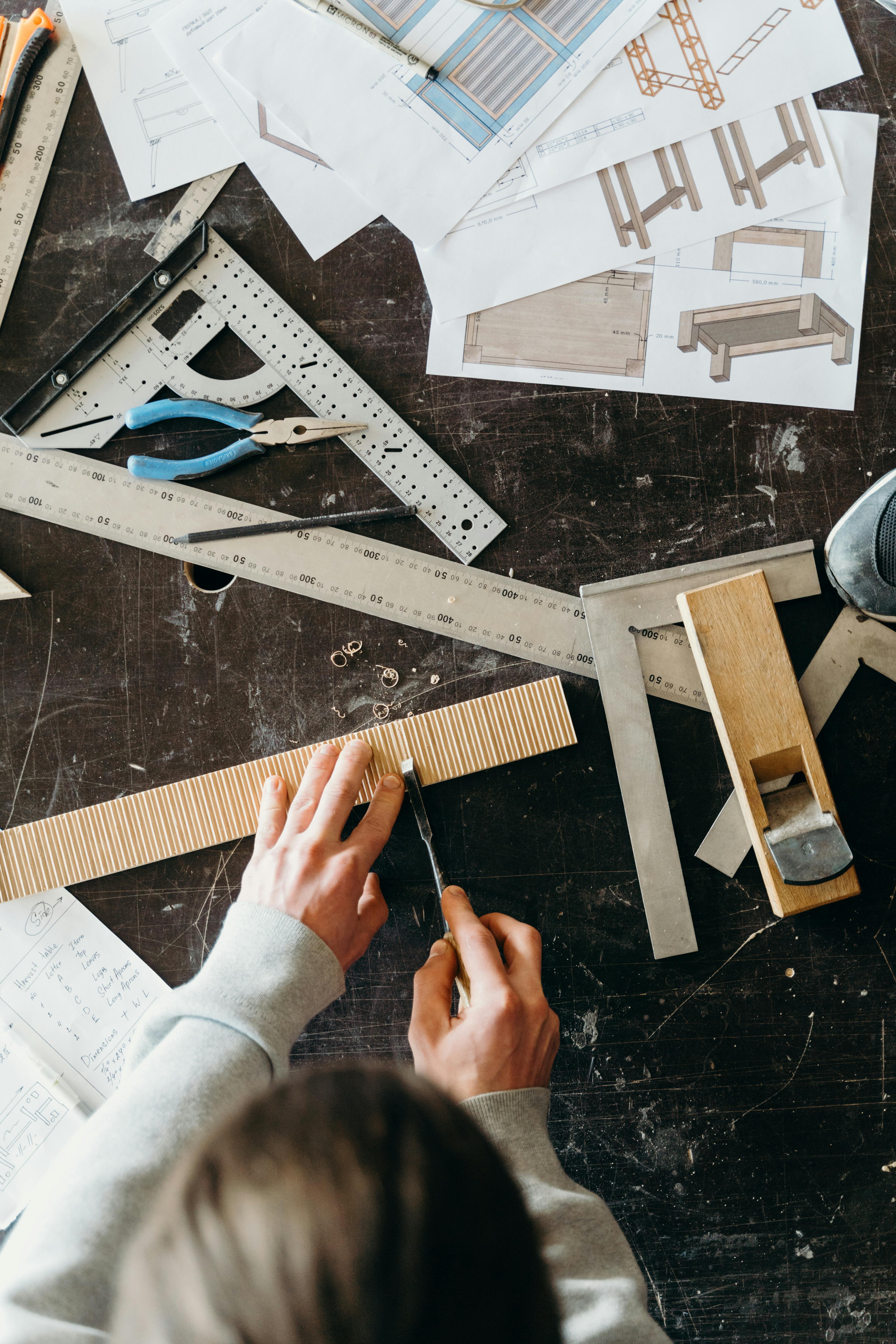 Person carving a piece of wood with a chisel on a workbench with carpentry tools and furniture sketches.